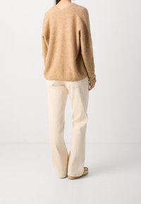Bežový vlněný svetr s kulatým zadním dílem a volným střihem, v kombinaci s krémovými širokými kalhotami a světlými hnědými botami.