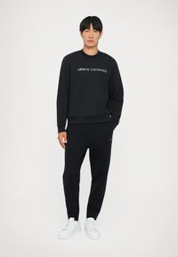 Melna džemperis ar "Armani Exchange" tekstu, apvienots ar melnām biksēm. Modelis valkā baltas kedas, stāvot pret vienkāršu fona.