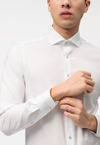 Weißes Hemd mit Knopfverschluss aus glattem Stoff. Verfügt über einen klassischen Kragen, lange Ärmel mit Manschetten und dezenten Knopfdetails nahe den Manschetten.