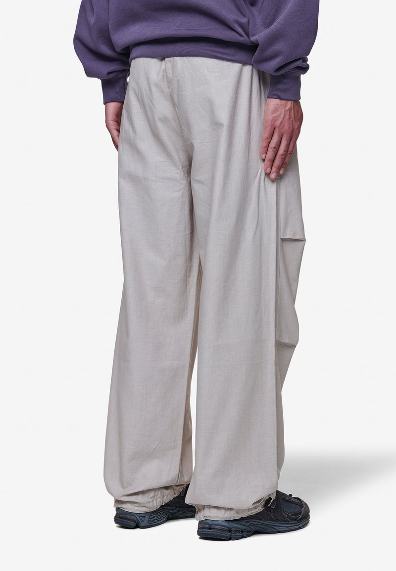 Jasno beżowe spodnie typu wide-leg mają luźny krój i teksturowany materiał, z bocznymi kieszeniami, połączone z ciemnymi sneakersami i fioletowym swetrem.