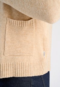 Beige stickad tröja med ribbad fåll och en fyrkantig ficka. Mjuk textur och subtilt fläckigt mönster. Etikett synlig på sidan.