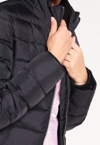 Czarna kurtka puchowa z wytłaczanym wzorem, zapięcie na zamek błyskawiczny i wysoki kołnierz. Wnętrze wykończone jest miękką podszewką.