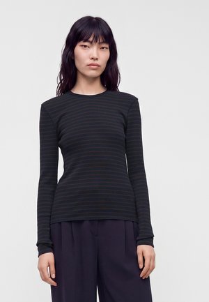 Femme aux longs cheveux foncés portant un t-shirt à manches longues rayé noir et marron et un pantalon ample foncé, debout devant un fond uni.