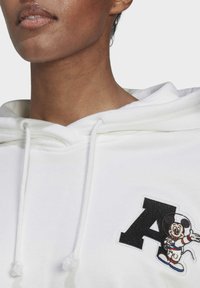 Sudadera blanca con cordones, que presenta un parche negro con una "A" y un personaje de dibujos animados bordado con una expresión sonriente. La tela parece suave.