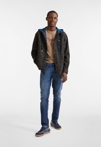 Chaqueta de cuadros oscuros con capucha azul, camiseta gráfica beige, jeans azules y zapatillas marinas con suelas marrones. Diseño moderno y sencillo.
