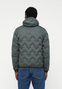 Puffer jacket in dark green with zigzag quilted pattern, hooded design, elastic cuffs, and a fitted hem. Worn with blue jeans.
