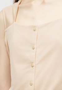 Haut beige à boutons avec bretelles fines porté sur une chemise beige transparente à manches longues, laissant apparaître le haut de la poitrine et les épaules.