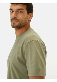 Oliven grøn t-shirt med korte ærmer i blød bomuld. Klassisk rund hals, tætsiddende pasform, med en lille logo detalje nær venstre ærme.