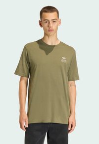 Bavlnené tričko olivovo zelenej farby s guľatým výstrihom, krátkymi rukávmi a bielym logom na hrudi. K tomu sú nosené tmavé rifle.