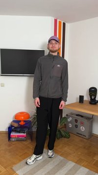 Jachetă gri cu fermoar și buzunar la piept, pantaloni negri și pantofi sport argintii. Se vede și o șapcă mov și tatuaje.