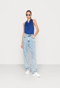 Top azul sem mangas com decote drapeado, combinado com jeans de perna larga em lavagens claras. Modelo usa óculos de sol redondos e tênis brancos.