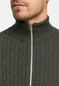 Mörkgrön, ribbad stickad tröja med hög krage och en silverfärgad dragkedja. Har ett flätmönster över hela tyget.