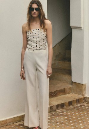 Femme portant des lunettes de soleil noires, un haut blanc transparent à pois, un pantalon blanc à jambes larges, debout près d'escaliers carrelés dans un environnement intérieur neutre.