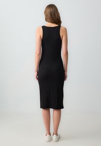Zwarte geribbelde midi-jurk met een getailleerde pasvorm, mouwloos ontwerp en minimale details. G gedragen met witte sneakers, gezien van achteren.