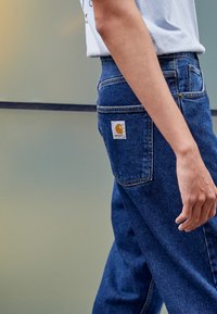 Oseba v modrih denim hlačah z logotipom Carhartt na zadnjem žepu in beli majici s kratkimi rokavi, v zamegljeni ozadju.