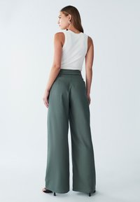 Pantalon large taille haute, léger, en tissu vert foncé, associé à un débardeur blanc côtelé, mettant en valeur une texture lisse.