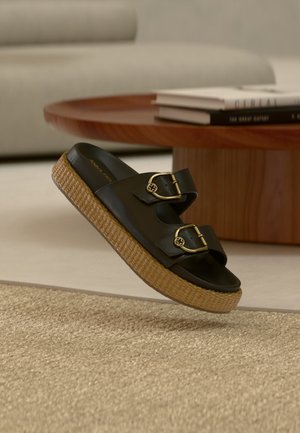 Svart slidesandal med to justerbare gullspenner og vevd platåsåle på beige teppe foran et trebord med bøker.