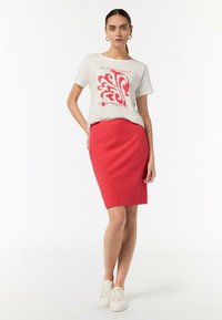 T-Shirt in Hellgrau mit rotem Grafiktext, kombiniert mit einem figurbetonten roten Rock. Modell trägt weiße Sneaker. Einfaches Design und glatte Textur.