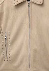 Giacca in suede finta beige con borchie argentate lungo il colletto e i lati, dotata di zip frontale e texture liscia.