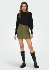 Haut noir à manches longues avec des manches texturées, associé à une mini jupe olive taille haute et des bottes noires brillantes jusqu'aux genoux.