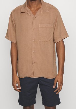 Mand iført en brun kortærmet skjorte med knapper og brystlomme samt sorte shorts til knæene mod en ensfarvet baggrund.