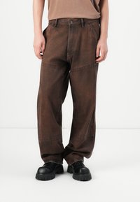 Persona con pantalones sueltos de color marrón oscuro con costuras visibles y zapatos negros de suela gruesa, de pie sobre un fondo blanco liso.