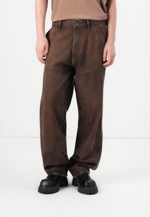 Person wearing loose, dark brown pants with visible seams and black chunky-soled shoes, standing on a plain white background.