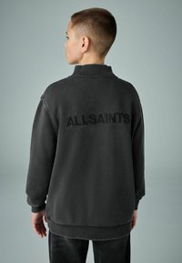 Tamsiai pilkas džemperis su aukšta apykakle, ant nugaros iškilmingas "ALLSAINTS" logotipas, pagamintas iš minkštos, tekstūruotos medžiagos.