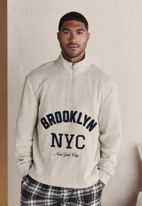 Fleece half-zip pullover i lys beige med "BROOKLYN NYC" tekst i marineblå. Har en blød tekstur og lange ærmer. Bæres med ternede pyjamabukser.