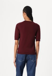 Burgundy neulepusero lyhyillä hihoilla, jossa on pyöreä pääntie ja ribbireuna, yhdistettynä korkeavyötäröisiin farkkuihin, nähtynä takaa.