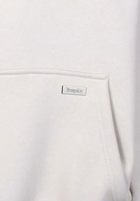 Helles beiges Sweatshirt mit einer Tasche auf der Vorderseite und einem metallischen Logo-Anhänger mit dem Text "Dropsix". Glatte Textur mit minimalistischen Design.