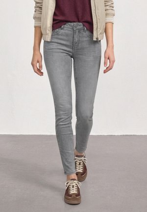 Person iført lysegrå skinny jeans, beige jakke, vinrød top og brune sneakers, der går på en grå gulv mod en hvid væg.