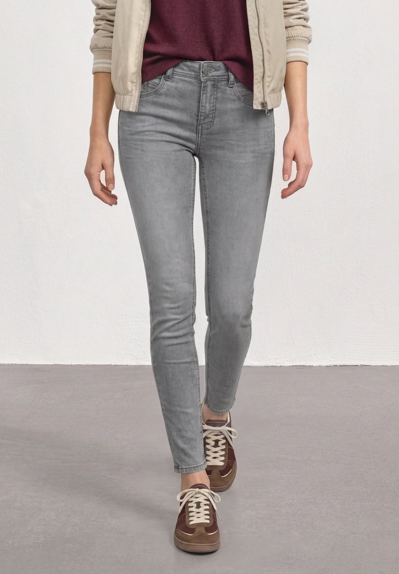 Personne portant un jean skinny gris clair, une veste beige, un haut bordeaux et des baskets marron marchant sur un sol gris devant un mur blanc.