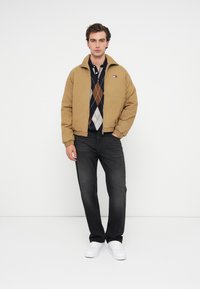 Καφέ κοντό bomber μπουφάν πάνω από πουλόβερ ναυτικού με σχέδιο argyle, συνδυασμένο με σκούρα τζιν και λευκά αθλητικά παπούτσια, σε λευκό φόντο.