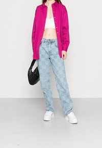 Kvinna som bär en lysande rosa skjorta med knappar över en vit croptop, ljusblå rutiga jeans, vita plattformssneakers, och håller en svart vadderad handväska.