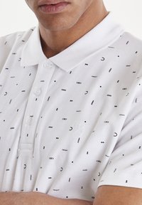 Bílá polo tričko s černými geometrickými vzory a textovými akcenty. Obsahuje límeček, tři knoflíky a krátké rukávy. Texturovaná látka.