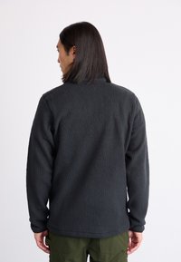 Zwarte fleece jacket met een gestructureerde oppervlakte, hoge kraag en lange mouwen, gedragen over groene broeken, gezien van achteren.