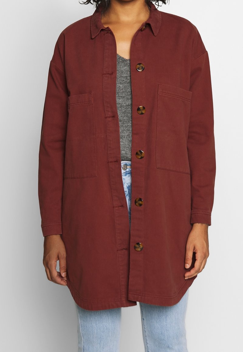 Veste boutonnée couleur bordeaux avec col, deux poches avant et boutons en écaille de tortue ; portée par-dessus un haut gris et un jean bleu clair.