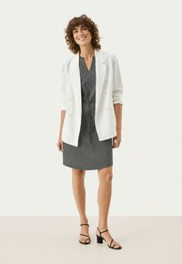 Vit linneblazer över en svartvit prickig klänning med åtdragen midja. Svarta remsandaler fulländar outfiten.