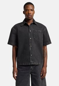Camicia nera in denim a maniche corte con colletto a punta, tasca frontale e bottoni argentati. Presenta una texture leggermente consumata.
