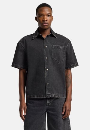 Black denim short-sleeve shirt with a pointed collar, front pocket, and silver buttons. Features a slightly distressed texture.