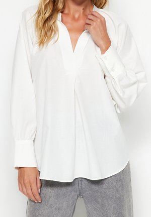 Femme portant un chemisier blanc ample à manches longues avec un col en V profond et des poignets boutonnés, associé à un jean gris délavé.