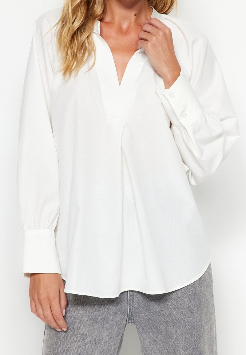 Femme portant un chemisier blanc ample à manches longues avec un col en V profond et des poignets boutonnés, associé à un jean gris délavé.