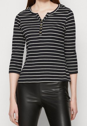 Femme portant un haut à manches trois-quarts à rayures horizontales noires et blanches avec des boutons au niveau de l'encolure et un pantalon en cuir noir.