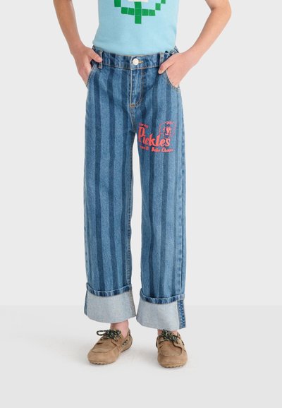 Enfant portant un jean à rayures verticales bleues avec des revers, un texte rouge « Pickles » et un graphique sur la cuisse, associé à des chaussures marron à lacets.