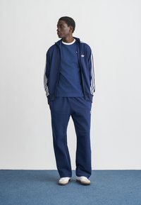 Navy tracksuit made of soft fabric, featuring white stripes on the jacket sleeves and logo. Pants have a relaxed fit and elastic waistband.