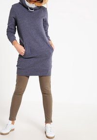 Vestido de sudadera gris marino con cuello en cowl, dos bolsillos frontales y forro a rayas. Combinado con leggings verde oliva y zapatillas blancas.