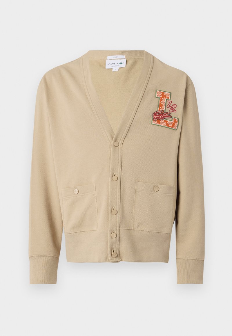 Cardigan beige en tissu doux avec un col en V, cinq boutons, deux poches avant et un écusson coloré brodé présentant un 'L' et un motif de crocodile.