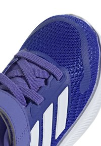 Zapatilla deportiva morada con parte superior de malla, acentos de panel texturizado, cordones planos y tres franjas blancas a lo largo del costado.