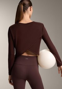 Nainen, jolla on poninhäntä ja ruskeaa urheiluvaatetusta, pitää valkoista teksturoitua lentopalloa lantiollaan, nähtynä takaa.